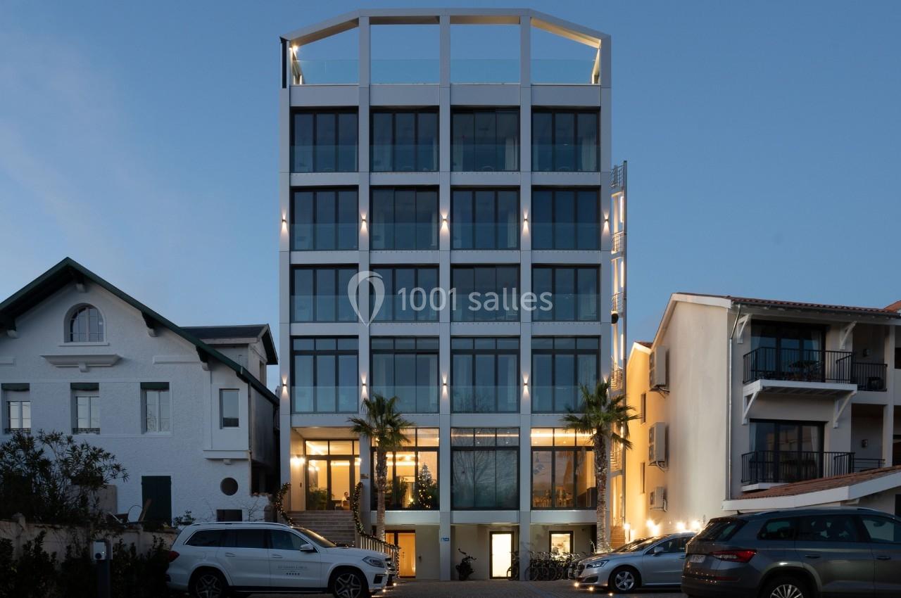 Façade d'un immeuble moderne à plusieurs étages, entouré de maisons et de voitures stationnées au crépuscule.