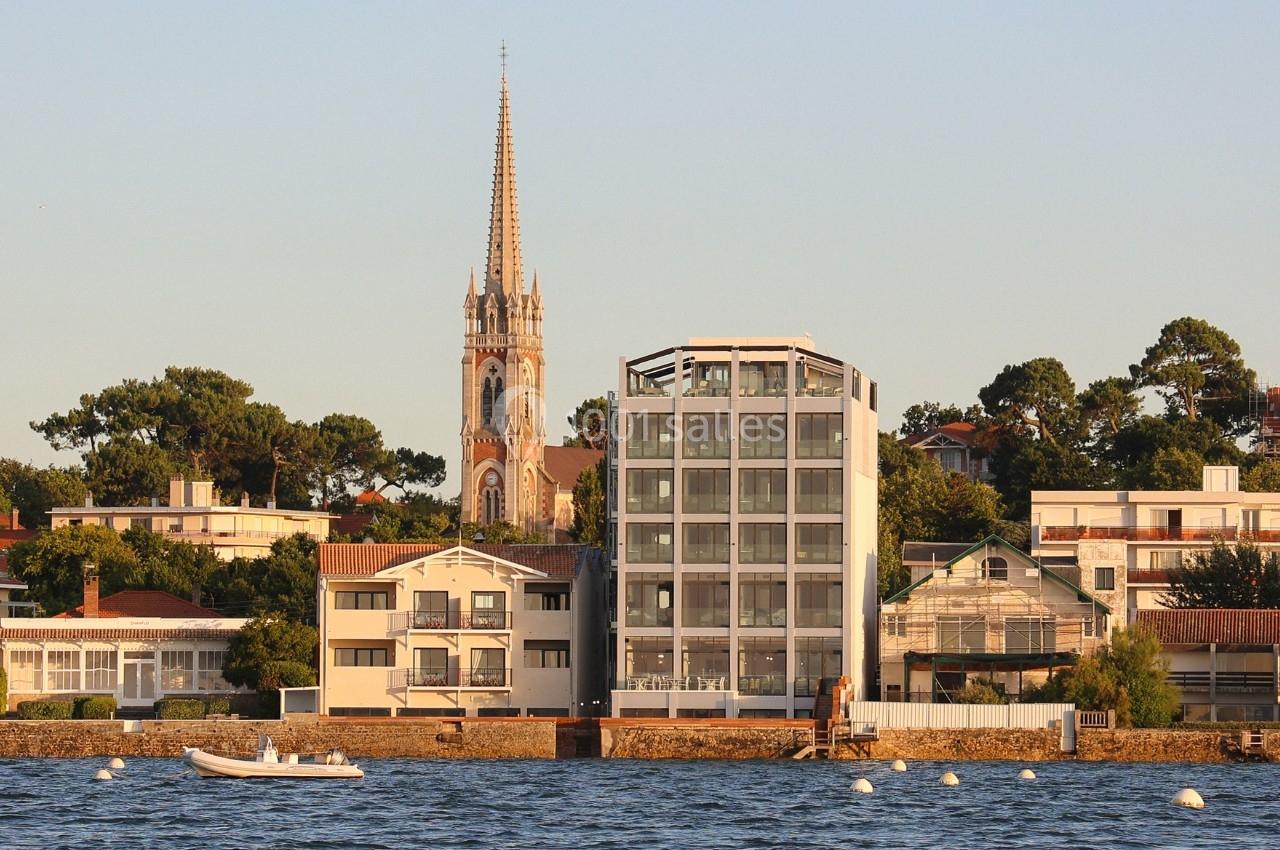 Vue d'un immeuble moderne et d'une église avec clocher, entourés de maisons, au bord d'une étendue d'eau.