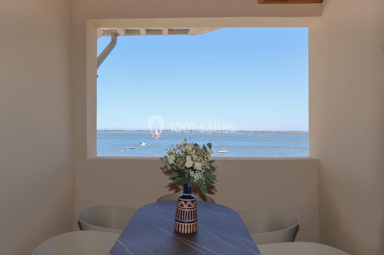Vue sur la mer à travers une fenêtre encadrant un vase avec des fleurs sur une table en pierre sombre.