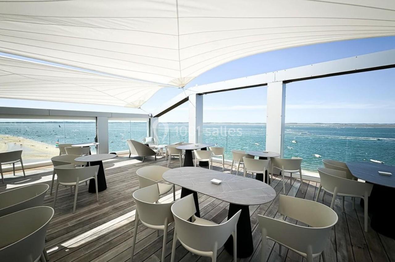 Terrasse en bois avec tables et chaises blanches, vue sur la mer et ciel dégagé, protégée par une toile tendue.