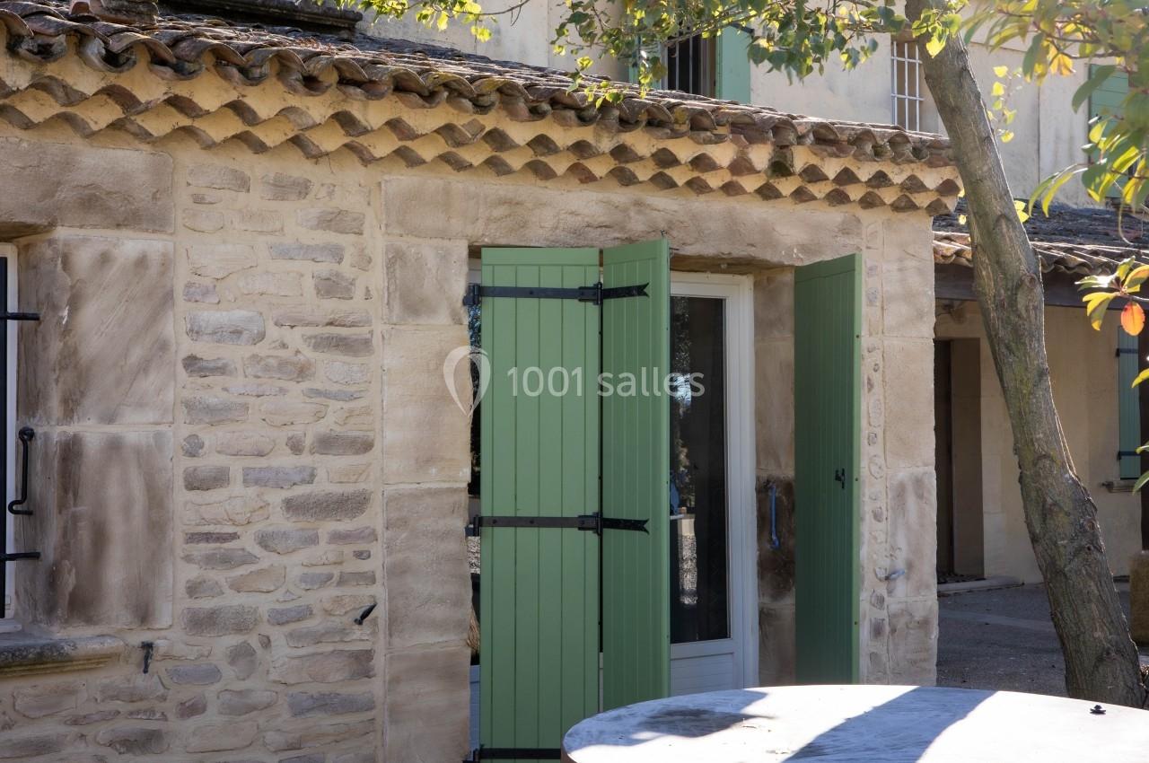Façade en pierre d'une maison avec volets verts ouverts, table en pierre au premier plan et arbre à droite.