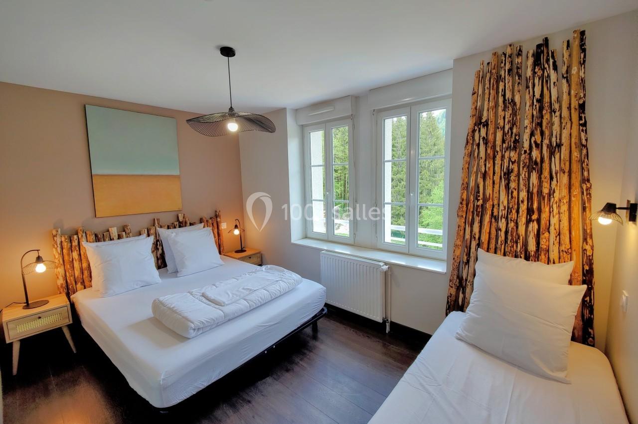Chambre lumineuse avec deux lits simples et un lit double, parquet sombre, grandes fenêtres et décoration naturelle.