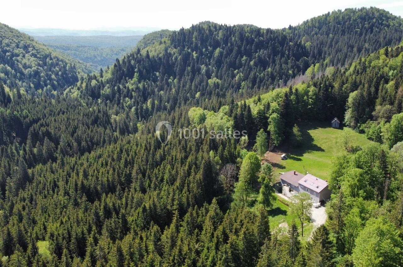 Vue aérienne d'une maison isolée entourée de forêts denses et de collines verdoyantes sous un ciel dégagé.