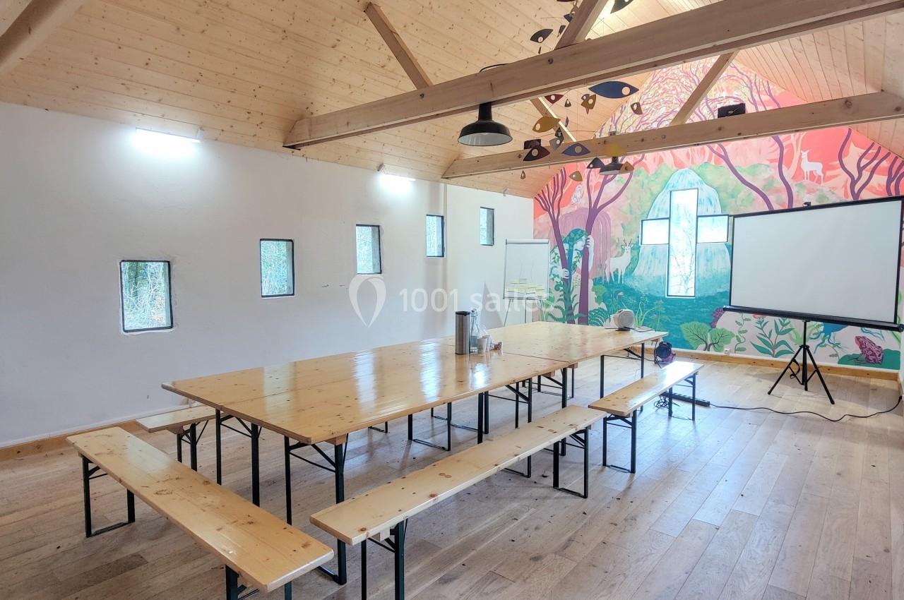 Salle lumineuse avec tables et bancs en bois, mur décoré d'une fresque colorée et écran de projection installé.