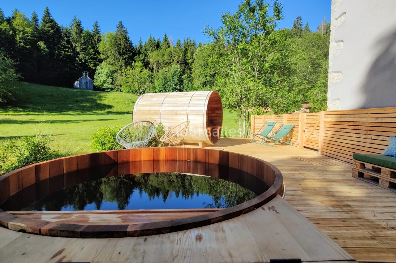 Terrasse en bois avec bain nordique, sauna en tonneau, chaises longues et vue sur une prairie bordée de forêt.
