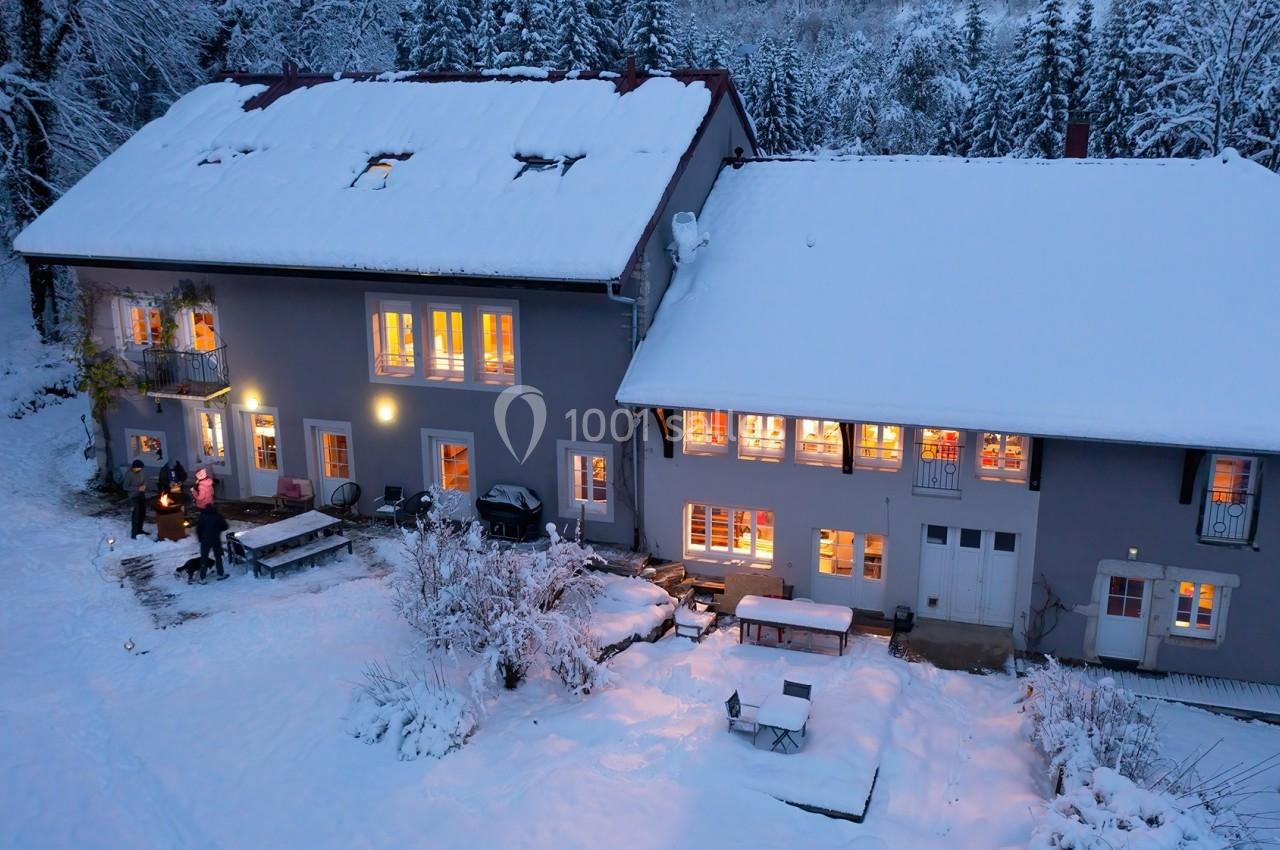 Vue aérienne d'une maison enneigée avec des lumières allumées, entourée d'arbres en hiver.