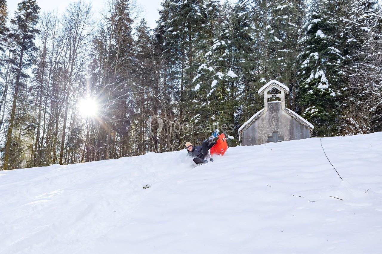 Une personne descend une pente enneigée sur une luge près d'une petite chapelle entourée d'arbres.