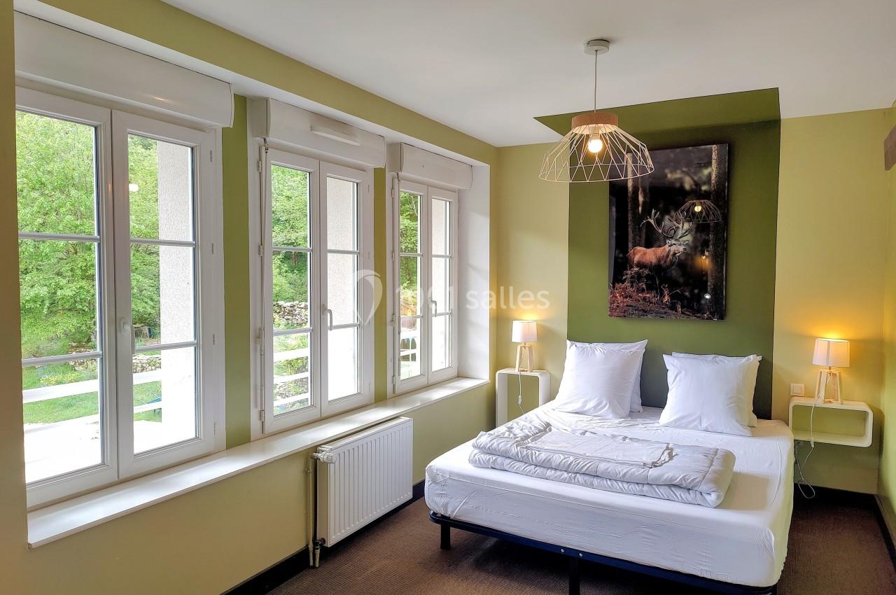 Chambre lumineuse avec lit double, grandes fenêtres donnant sur un paysage verdoyant et décor minimaliste.