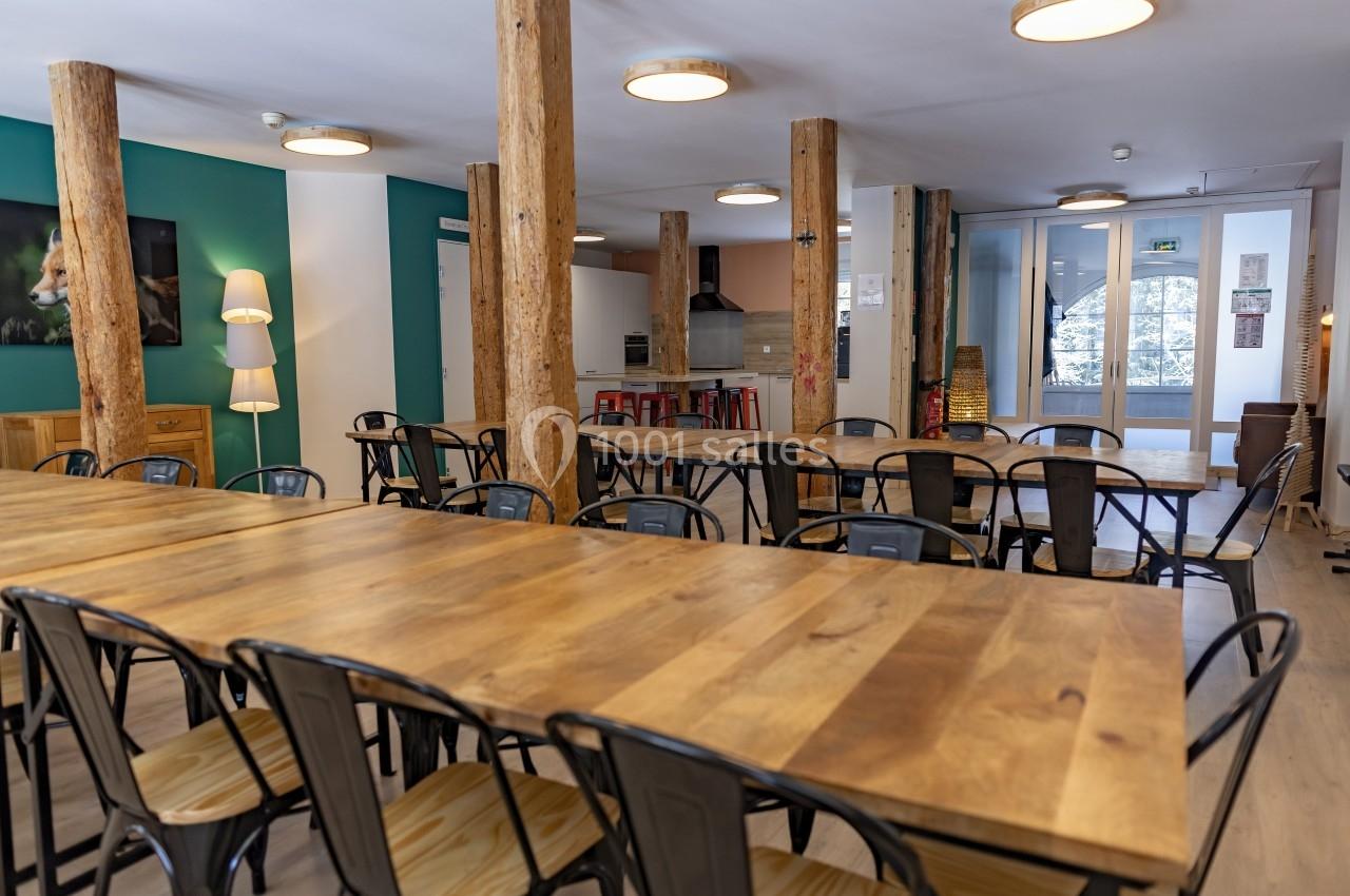 Salle lumineuse avec de grandes tables en bois, chaises métalliques, colonnes en bois et cuisine ouverte en arrière-plan.