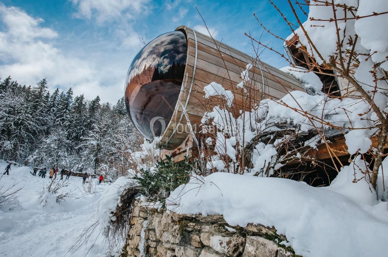 Cabane en bois avec une grande fenêtre arrondie, entourée de neige et de végétation hivernale, près d'une forêt.