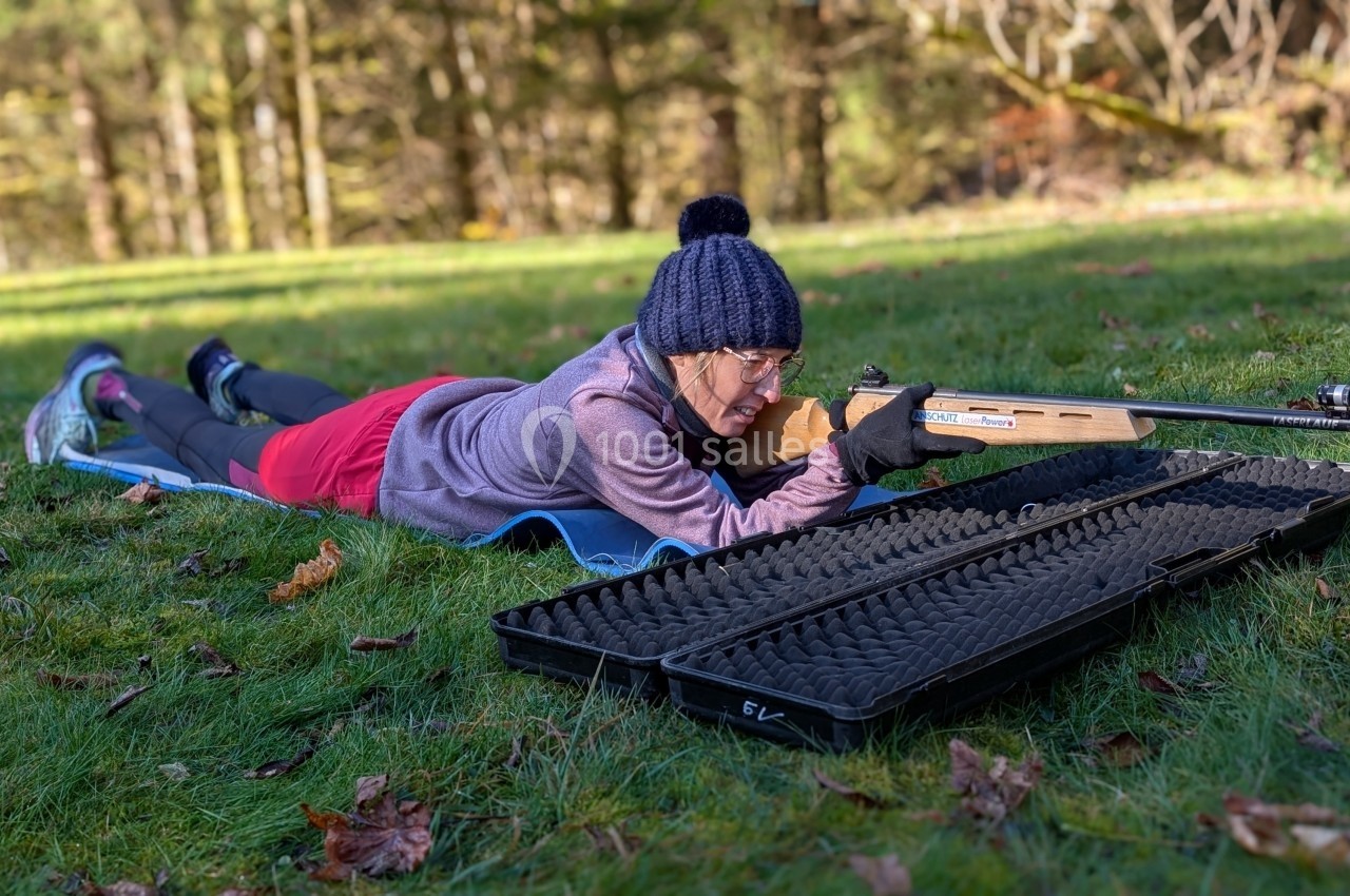 Une personne allongée sur l'herbe vise avec une carabine, un étui ouvert posé à côté, dans un environnement boisé.
