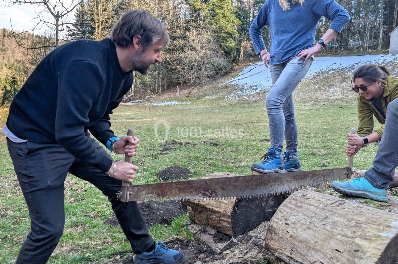 Deux personnes scient un tronc d’arbre avec une grande scie, sous le regard de deux autres dans un cadre naturel.