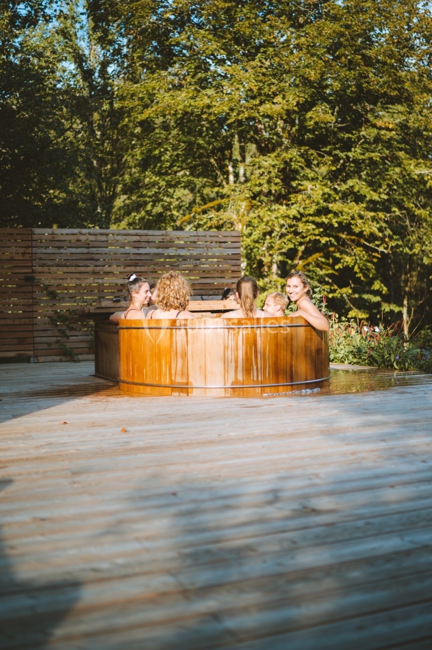 Des personnes se détendent dans un bain nordique en bois, entouré de végétation et d'une terrasse en bois.