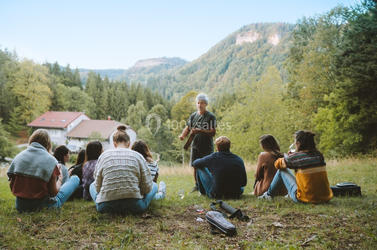 Un groupe de personnes assises en cercle dans une clairière, écoutant une personne debout devant un paysage boisé.
