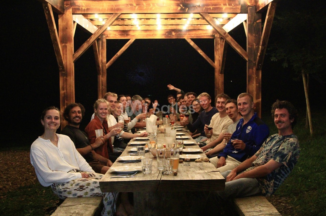Un groupe de personnes assises autour d'une longue table en bois sous une pergola éclairée, de nuit.