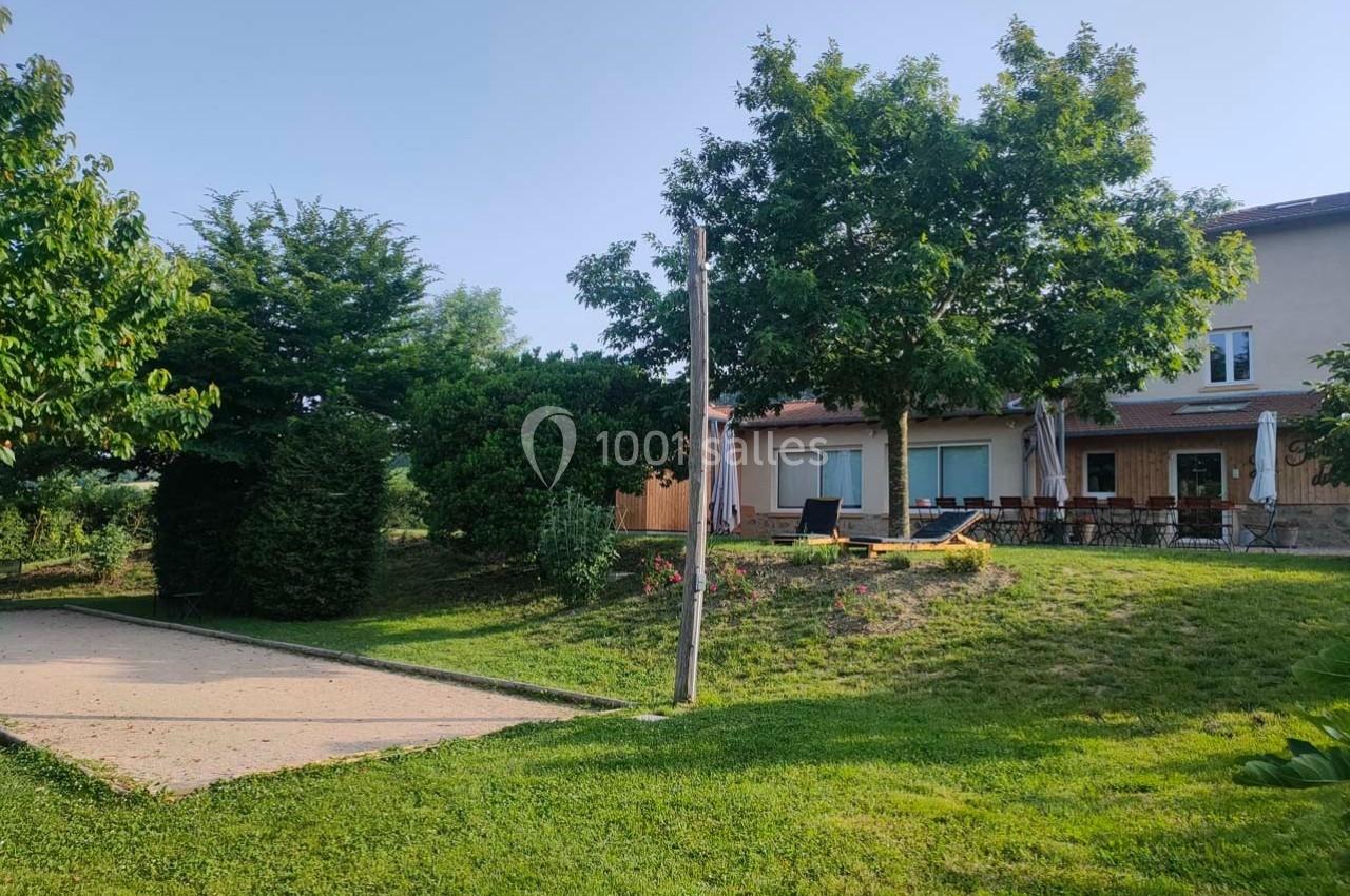 Terrain de pétanque ombragé devant une maison avec terrasse, entouré de pelouse et d'arbres.