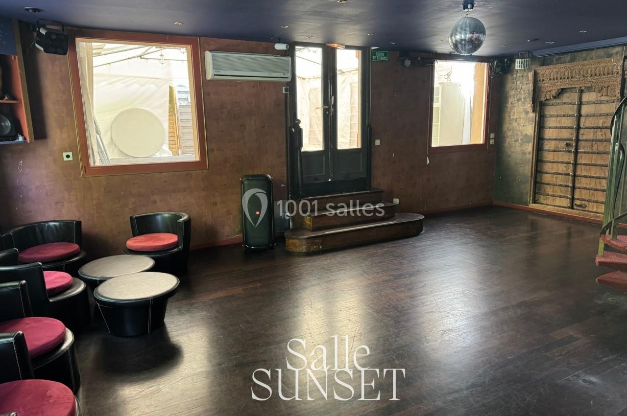 Salle vide avec parquet sombre, murs en bois, fauteuils noirs, tables basses, et une boule disco suspendue au plafond.