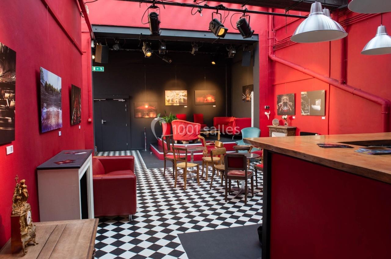 Salle avec murs rouges, sol en damier noir et blanc, mobilier varié et scène en fond éclairée par des projecteurs.