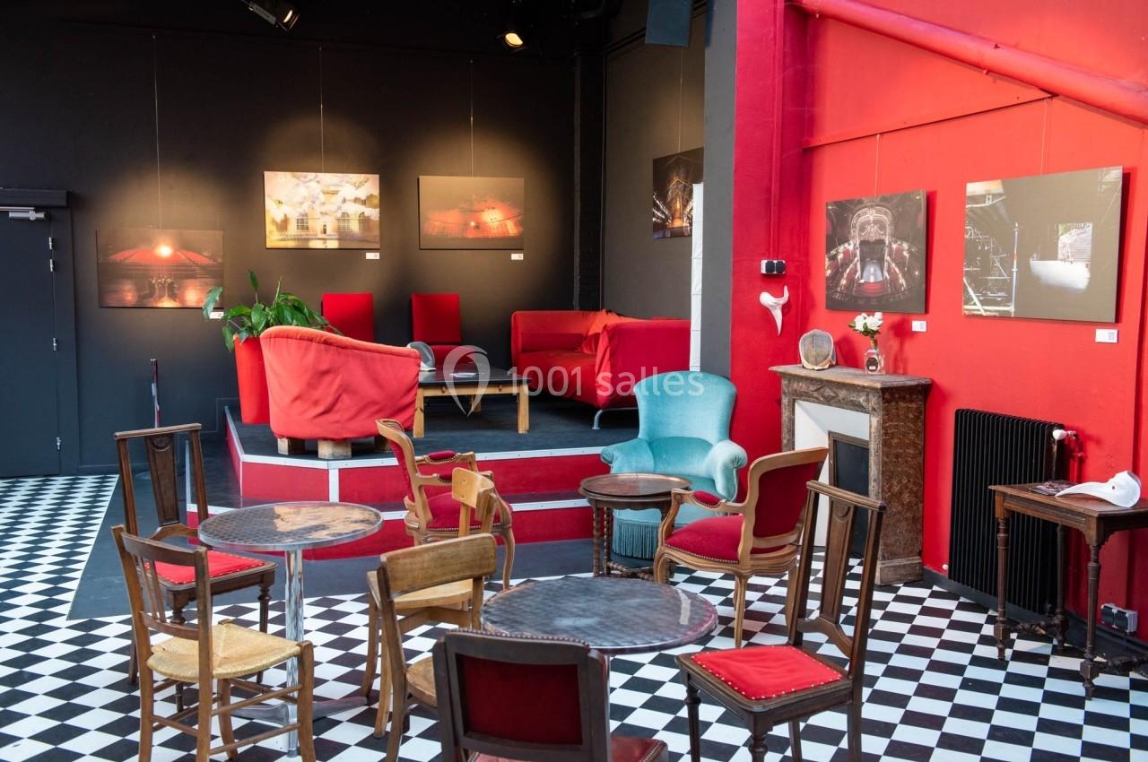 Espace intérieur avec tables, chaises variées, canapé rouge, murs noirs décorés de tableaux et sol en damier noir et blanc.