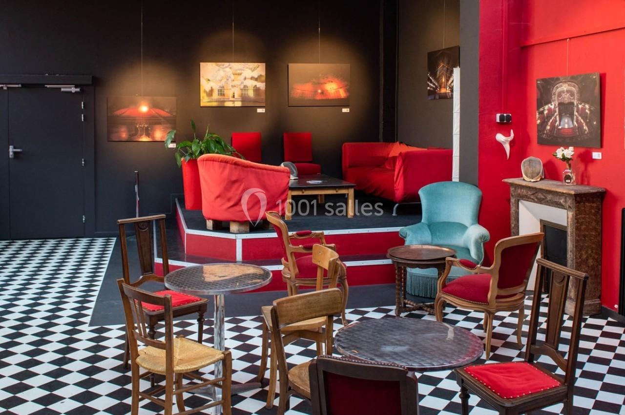 Salle avec tables et chaises en bois, fauteuils rouges et verts, sol en damier noir et blanc, et tableaux accrochés au mur.