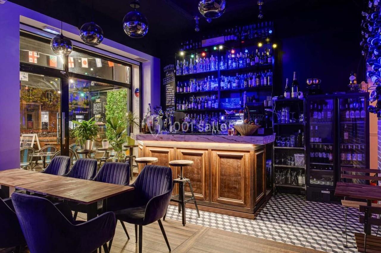 Intérieur d'un bar avec un comptoir en bois, des étagères éclairées de bouteilles et des tables entourées de chaises.