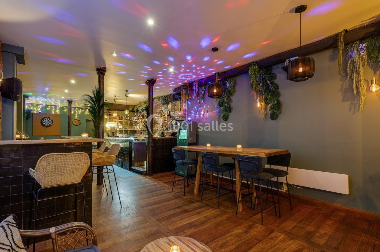 Intérieur d'un bar avec éclairage tamisé, décorations végétales, tables en bois et lumières colorées au plafond.