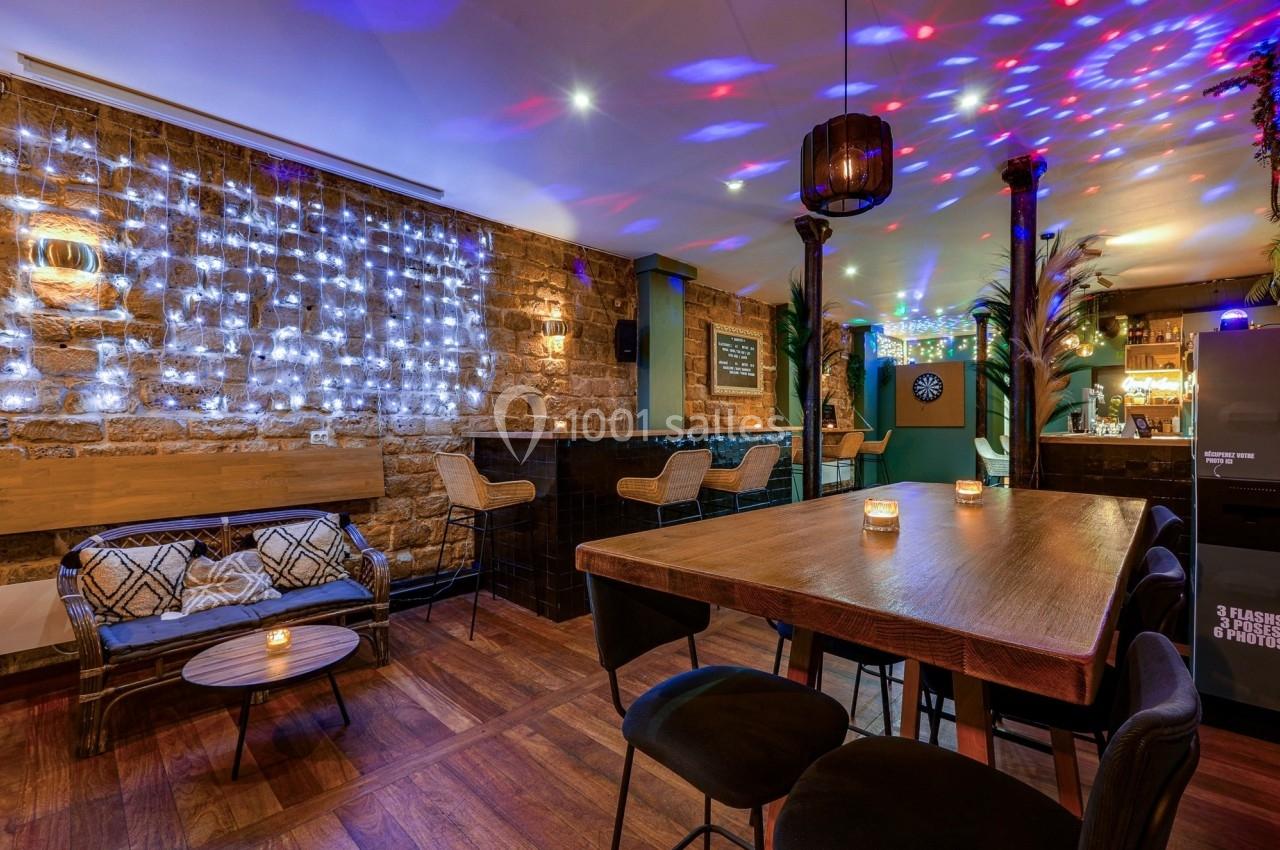 Salle de bar chaleureuse avec murs en pierre, guirlandes lumineuses, table haute en bois et éclairage coloré.
