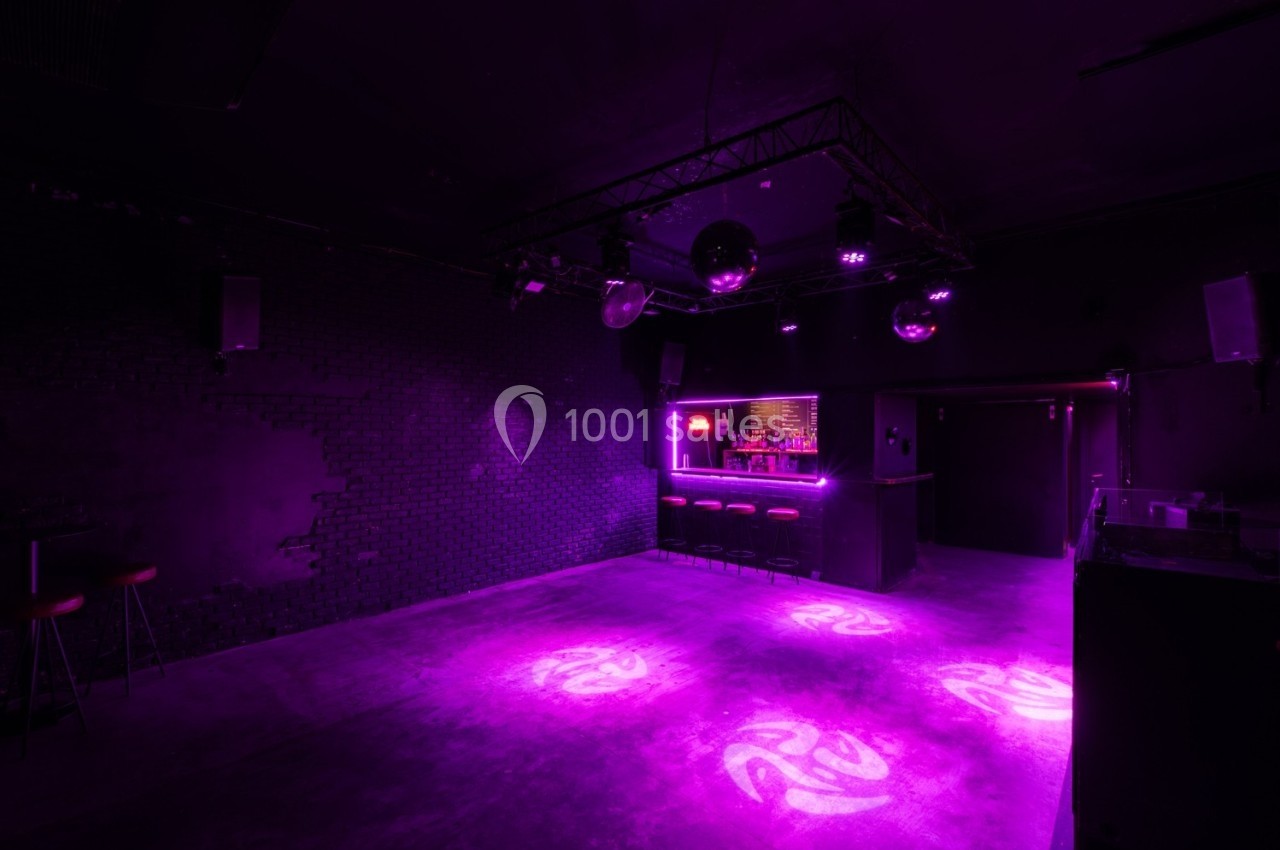 Salle de danse vide avec éclairage violet, bar illuminé en arrière-plan et murs sombres.
