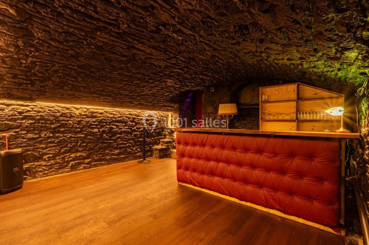 Cave voûtée en pierre avec un bar en cuir rouge, éclairage chaleureux et étagères en bois à l'arrière.
