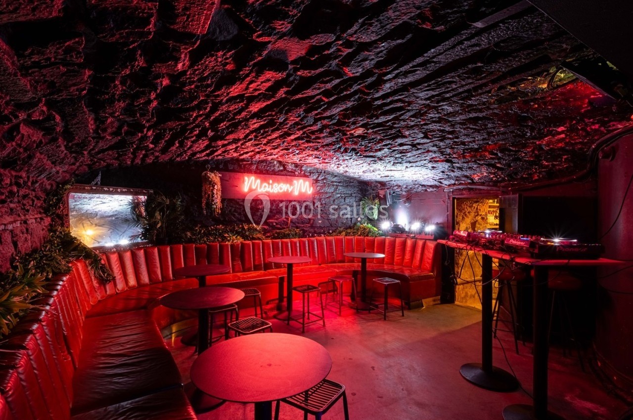 Salle voûtée en pierre avec banquettes rouges, tables basses, éclairage néon et espace DJ équipé.