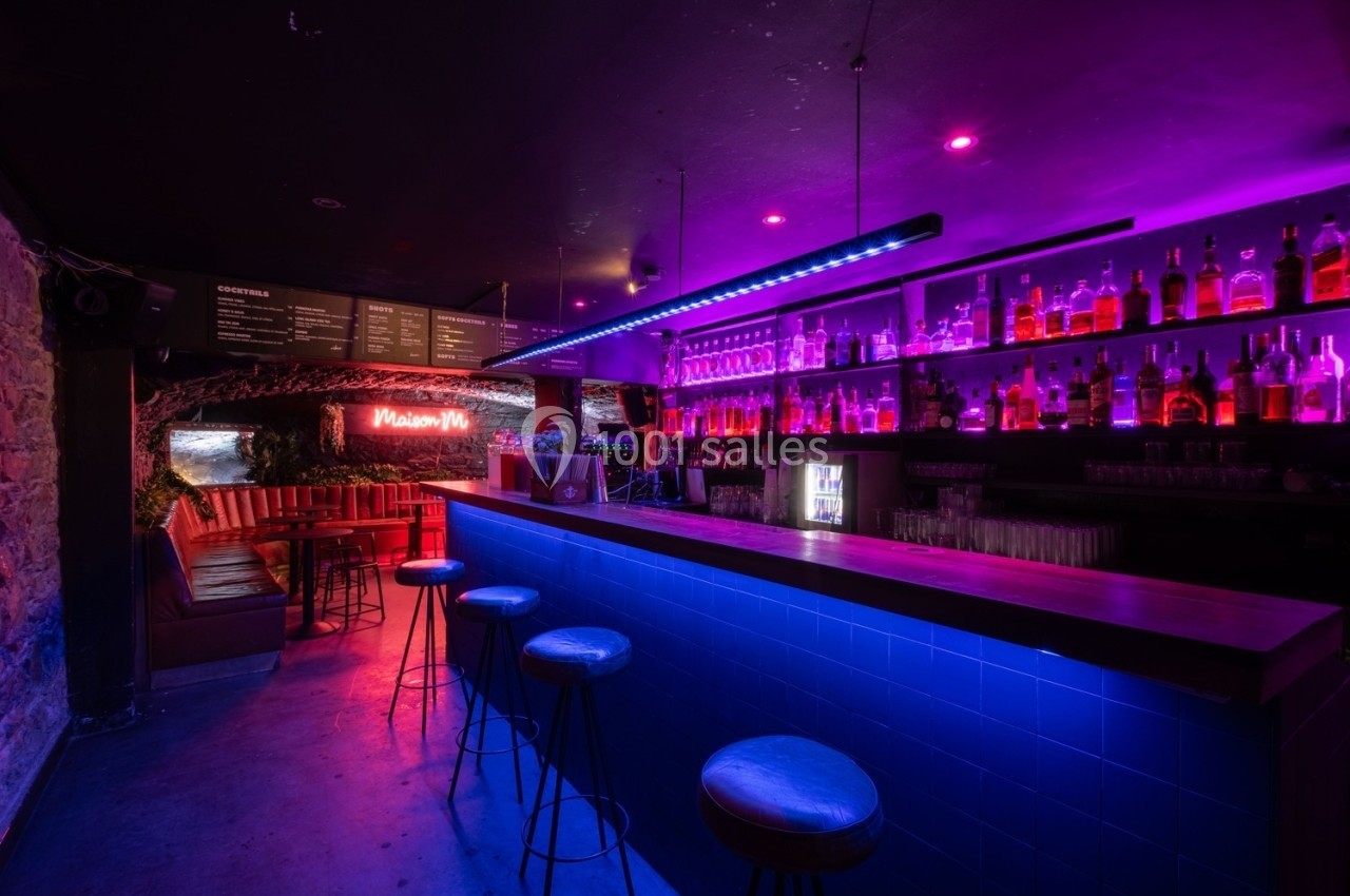 Bar avec comptoir éclairé en bleu, étagères de bouteilles illuminées en rose et coin banquette en arrière-plan.