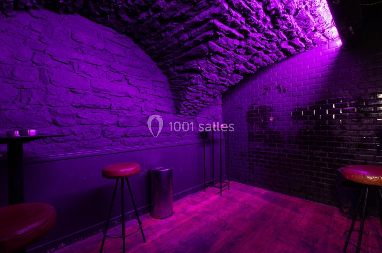 Salle voûtée en pierre avec éclairage violet, tabourets hauts, tables et sol en bois.