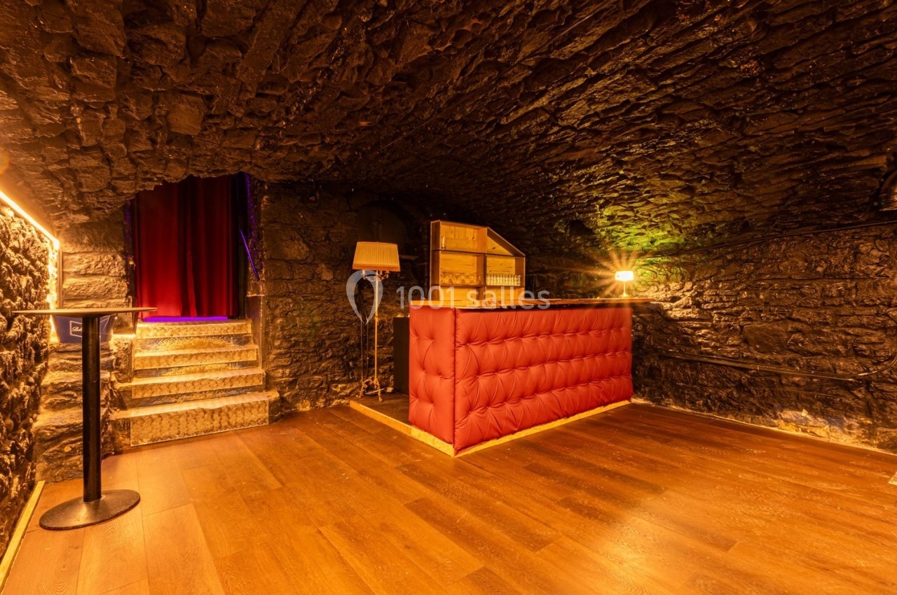 Salle voûtée en pierre avec un comptoir rembourré rouge, éclairage chaleureux et escalier menant à une entrée.
