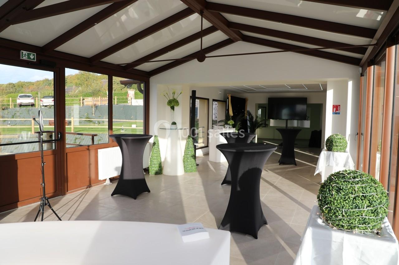 Salle lumineuse avec tables hautes noires, décorations végétales et grandes baies vitrées donnant sur l'extérieur.