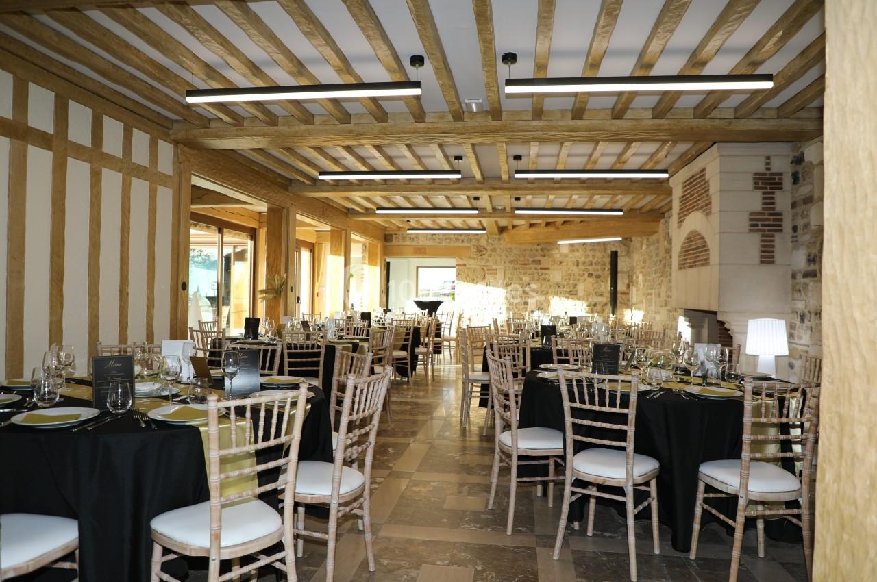 Salle de réception lumineuse avec tables dressées, chaises en bois clair et poutres apparentes au plafond.