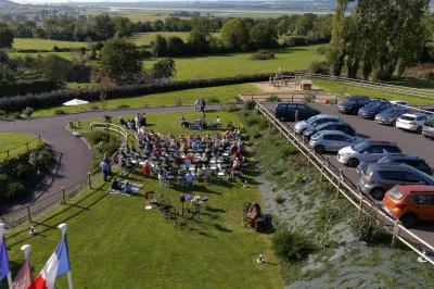 Aire de jeux en bois sur sol sablonneux, entourée de verdure et située près de bâtiments ruraux sous un ciel bleu.