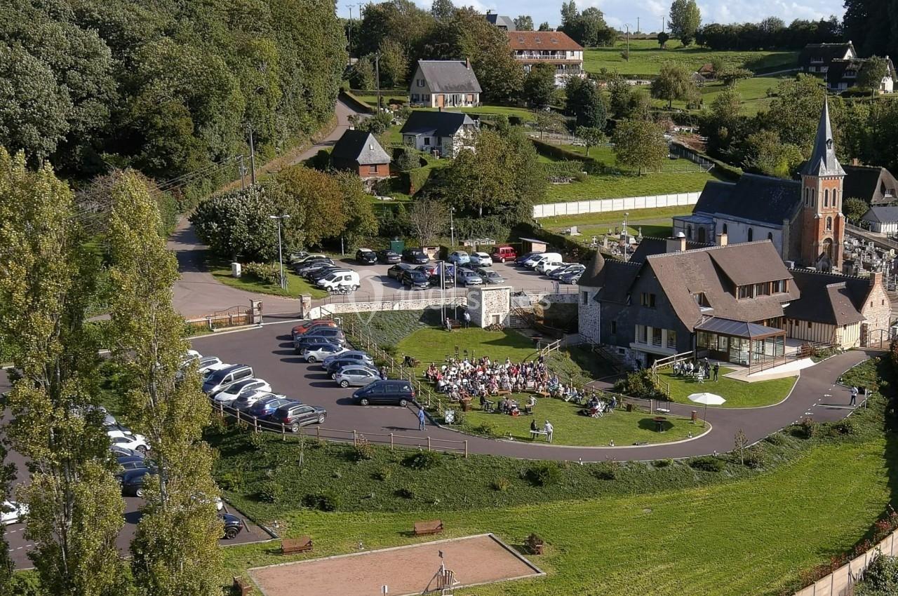 Vue aérienne d'un village avec une église, un parking, une aire de jeux et des personnes rassemblées sur une pelouse.
