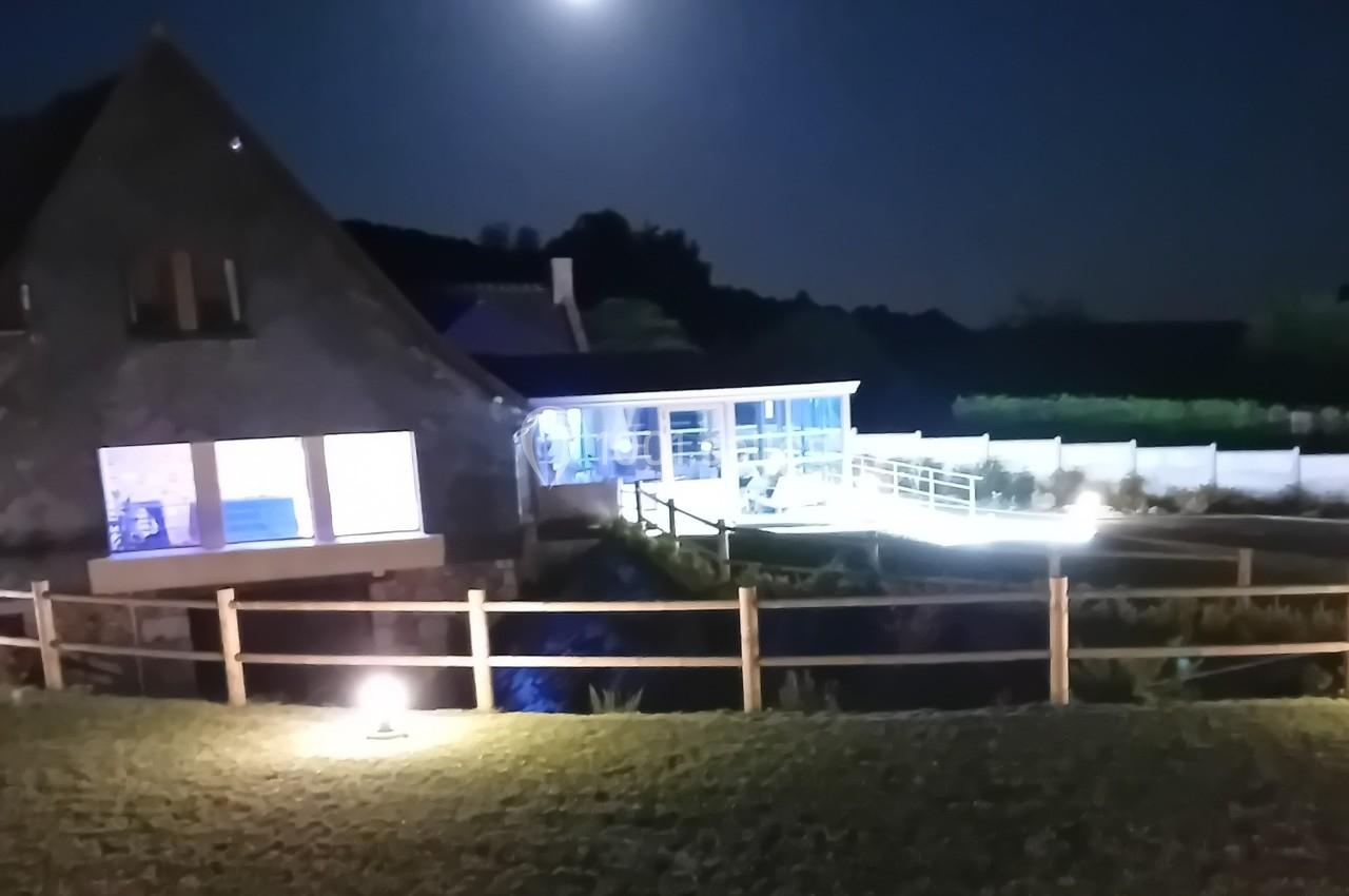 Maison avec extension moderne éclairée la nuit, entourée d'une clôture en bois et d'un jardin.