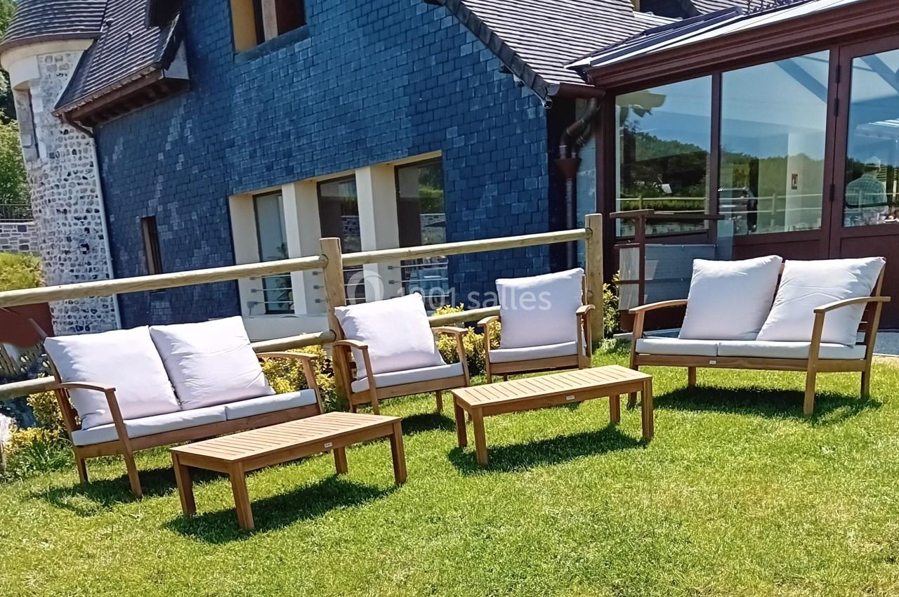 Mobilier de jardin en bois avec coussins blancs disposé sur une pelouse devant une maison en pierre et verre.