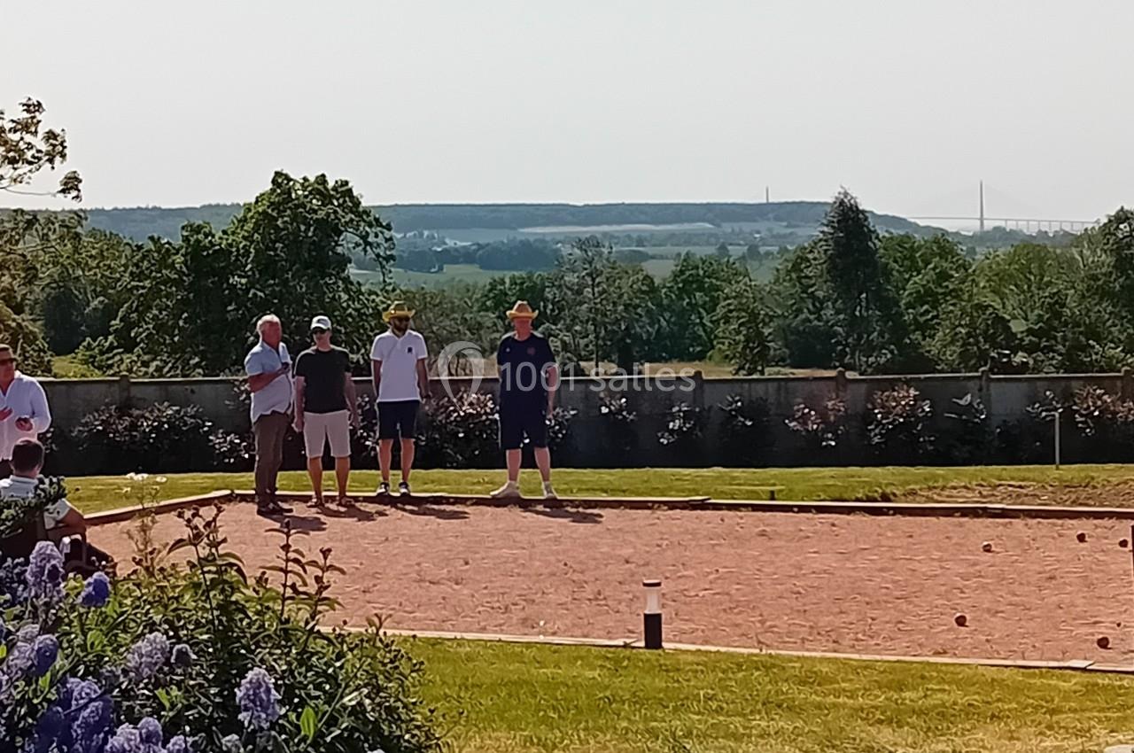 Des personnes jouent à la pétanque sur un terrain ensoleillé, avec un paysage verdoyant et un pont visible au loin.