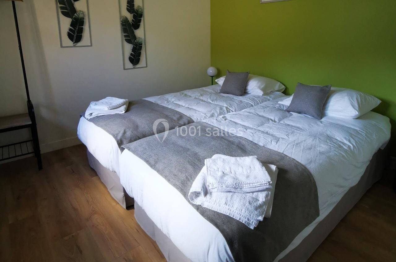 Deux lits simples avec draps blancs, couvertures grises et serviettes pliées, dans une chambre au mur vert.