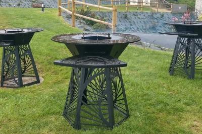 Aire de jeux en bois sur sol sablonneux, entourée de verdure et située près de bâtiments ruraux sous un ciel bleu.