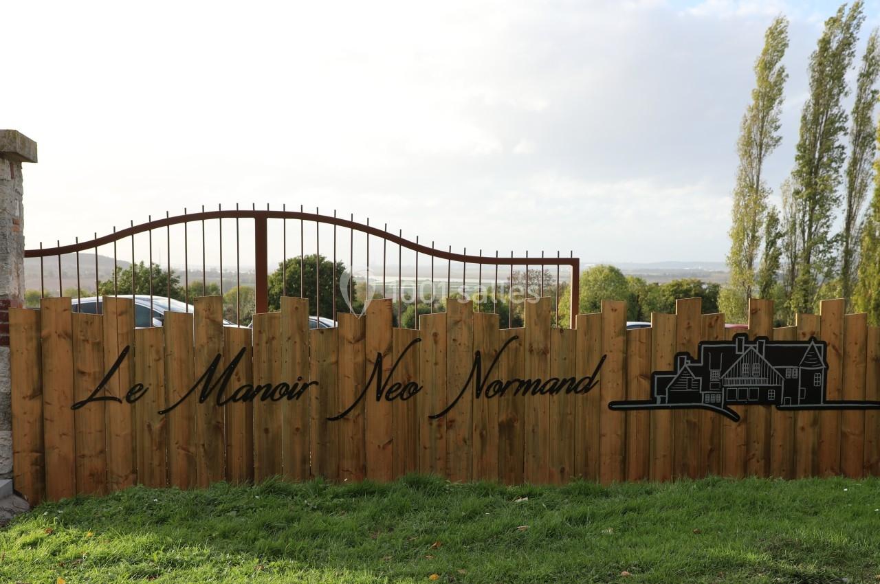 Clôture en bois avec le texte ’Le Manoir Néo Normand’ et un dessin de manoir, sur fond de paysage rural.