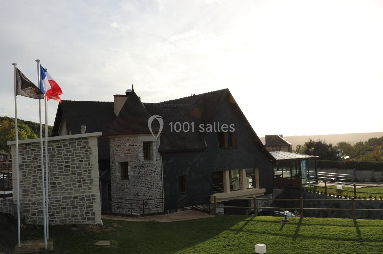 Bâtiment en pierre et ardoise avec des drapeaux français et régional, entouré de verdure sous un ciel clair.