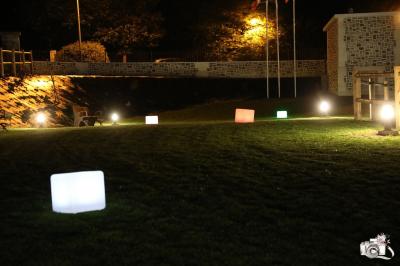 Aire de jeux en bois sur sol sablonneux, entourée de verdure et située près de bâtiments ruraux sous un ciel bleu.
