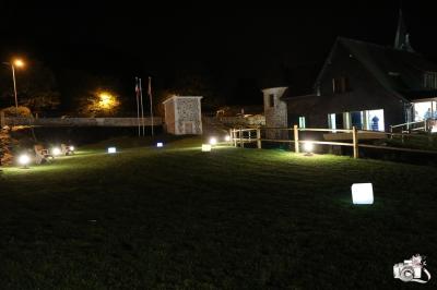 Aire de jeux en bois sur sol sablonneux, entourée de verdure et située près de bâtiments ruraux sous un ciel bleu.