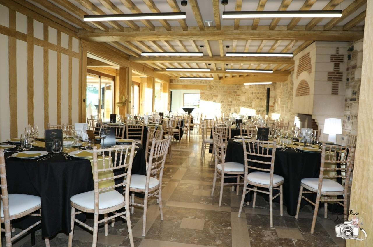 Salle de réception lumineuse avec tables dressées, nappes noires et chaises en bois, dans un décor rustique en pierre et…