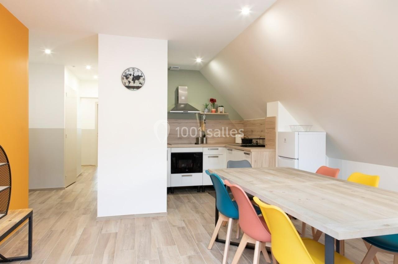 Salle à manger lumineuse avec table en bois, chaises colorées et cuisine ouverte équipée en arrière-plan.