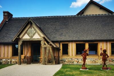 Aire de jeux en bois sur sol sablonneux, entourée de verdure et située près de bâtiments ruraux sous un ciel bleu.
