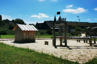 Aire de jeux en bois sur sol sablonneux, entourée de verdure et située près de bâtiments ruraux sous un ciel bleu.